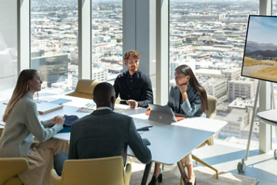 Four people in a meeting at a desk in front of a large wall of windows overlooking a city.