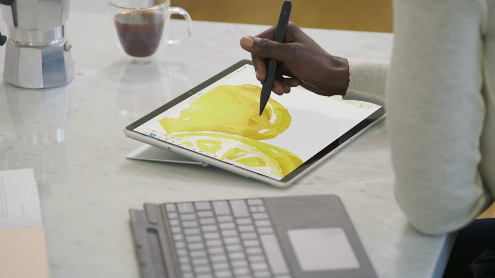 スリム ペンを使用して、Surface Pro 8 の画面上の黄色の画像に色を追加し、近くに取り外し可能なタイプ カバー キーボードがあるユーザー。