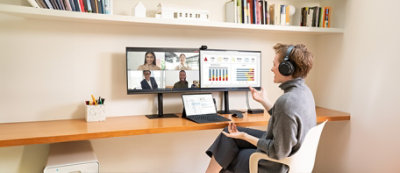 A person in headphones sits at a desk with two monitors; one screen shows a video conference with four participants