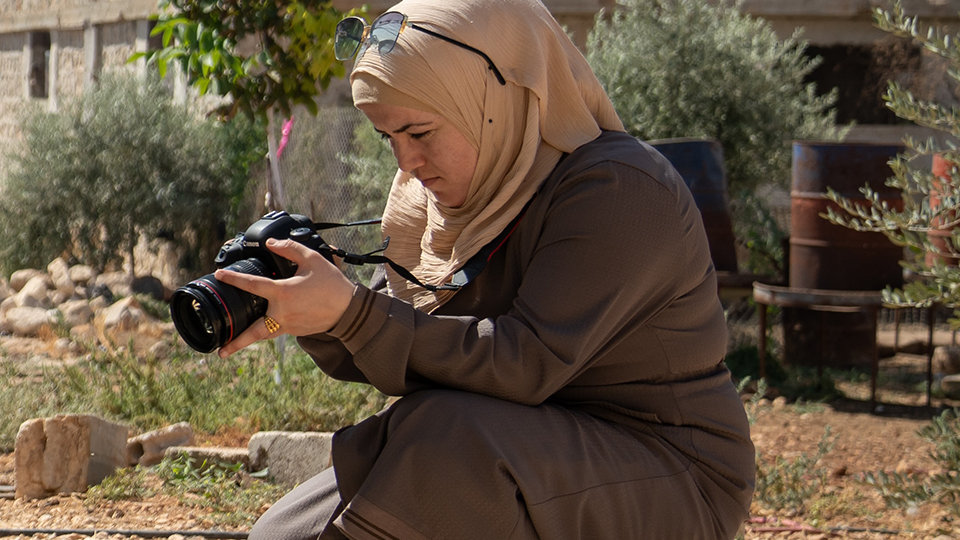 A woman in a hijab crouches outdoors while looking at a camera.