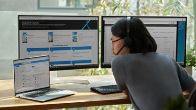 A person wearing a headset works at a desk with two monitors and a laptop.