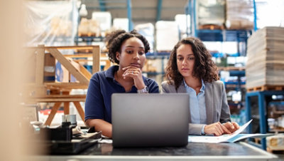 Female worker with supervisor in a warehouse working on laptop.