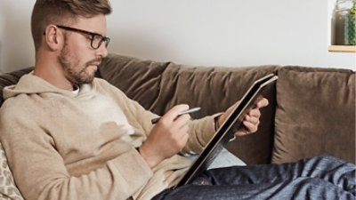 Una persona sdraiata su un divano che usa una penna e un tablet.