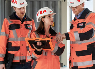 Trois employés de Strabag portant des casques de protection et des vêtements réfléchissants. Ils discutent et regardent une tablette