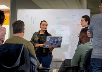 Person presenting to a group in a meeting room, standing in front of a whiteboard holding a laptop.