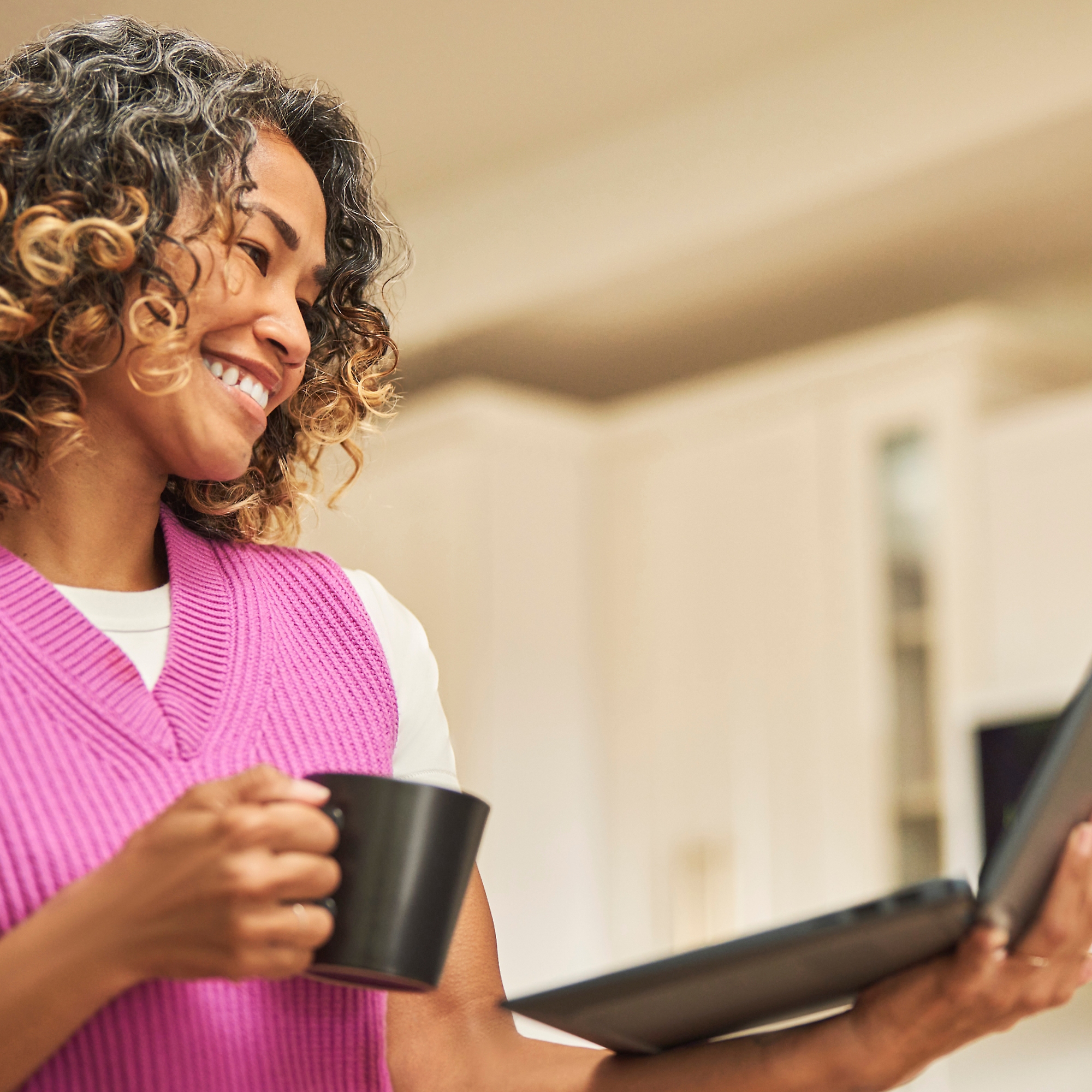 Een vrouw die glimlacht met een koffiekop in de ene hand en een laptop in de andere