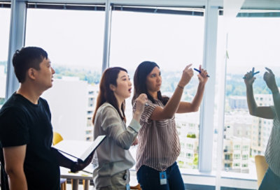 صورة لثلاثة أشخاص يقفون أمام لوح معلومات ويتناقشون. أحدهما يحمل جهاز كمبيوتر محمول.