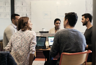 Ảnh năm người đang họp tại bàn tròn. Họ đều có máy tính xách tay. Có một bảng trắng với các ghi chú trong nền.