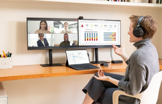自宅オフィスの机に座り、Microsoft Teams の通話と Dynamics 365 を表示する 2 台の外付けモニターに接続された、Outlook を表示する Surface Pro 9 で作業する女性。
