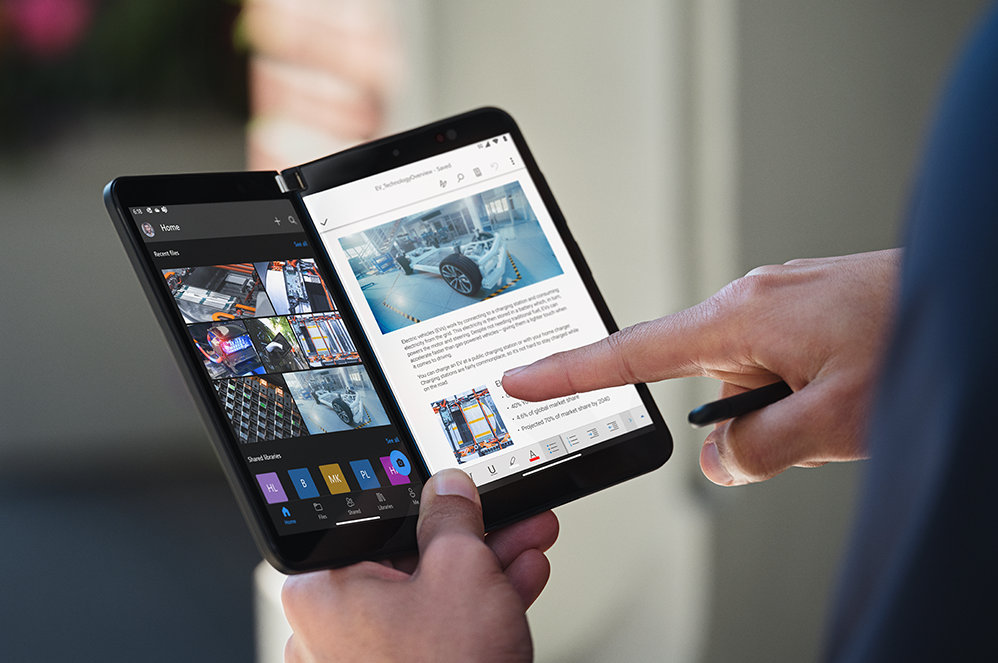 Iemands hand houdt een Surface Duo voor zakelijk gebruik vast alsof het een boek is en wijst met een vinger naar het scherm.