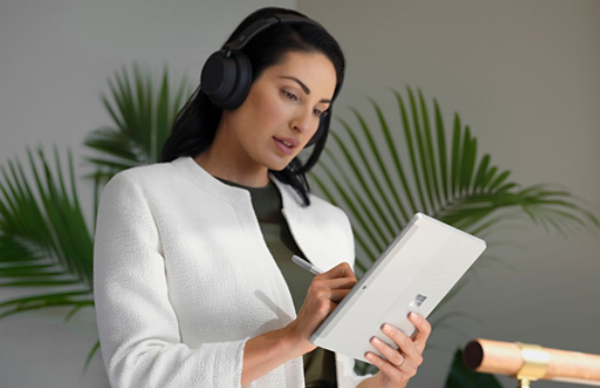 A person wearing Surface Headphones 2+ using Surface Pen to navigate Surface Go 4 in tablet mode at work