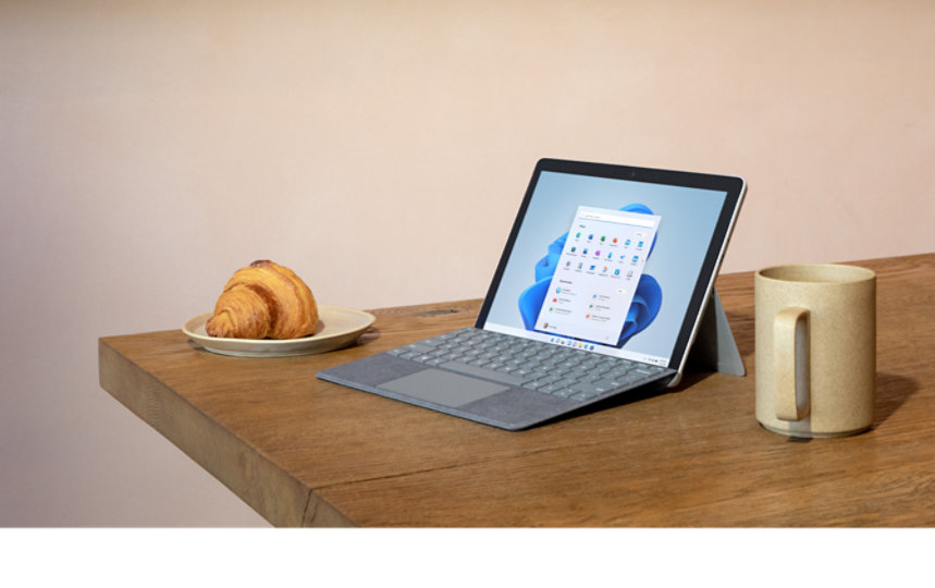 Surface Go 3 com uma capa teclado acoplada pousada numa mesa, e rodeada por um croissant e uma chávena de café.