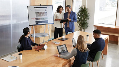 A woman draws on Surface Hub in an office meeting with several people around a table.