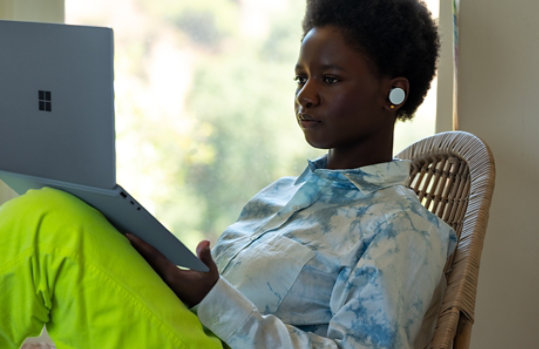 Une femme assise sur une chaise et portant des écouteurs utilise son Surface Laptop Go posé sur son genou.