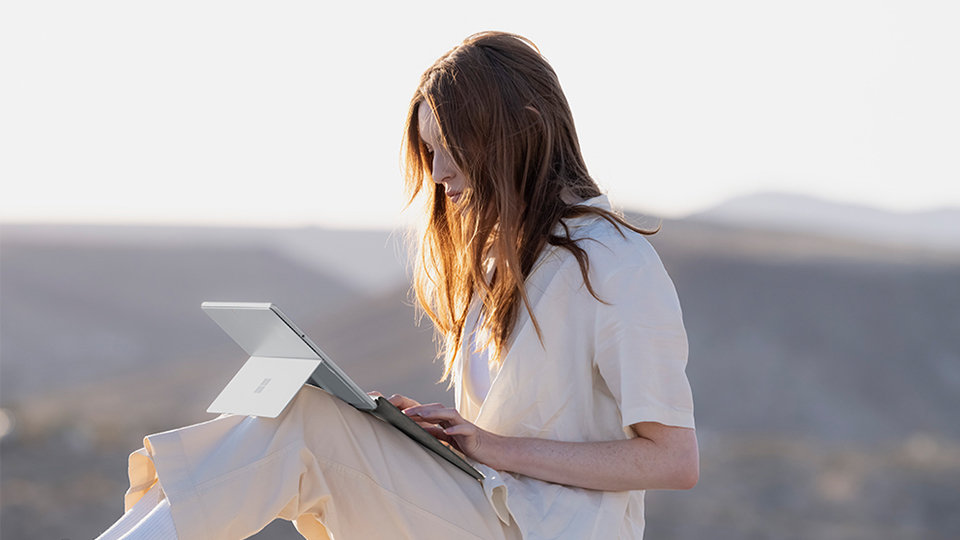 Eine Person sitzt in der Sonne in der Nähe von Bergen und tippt auf einem Surface Pro Signature Keyboard mit Fingerabdruckleser.