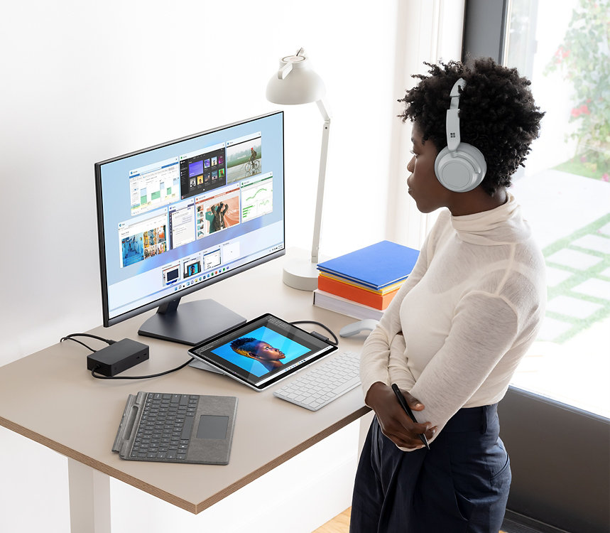 Eine Person, die Surface Headphones trägt, steht vor einem Schreibtisch mit einem Surface-Gerät, Type Cover und Monitor darauf.