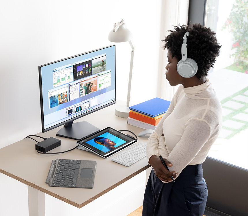 A woman wearing Surface headphones at a standing desk with Surface Pro X docked with a monitor and two keybaords nearby.