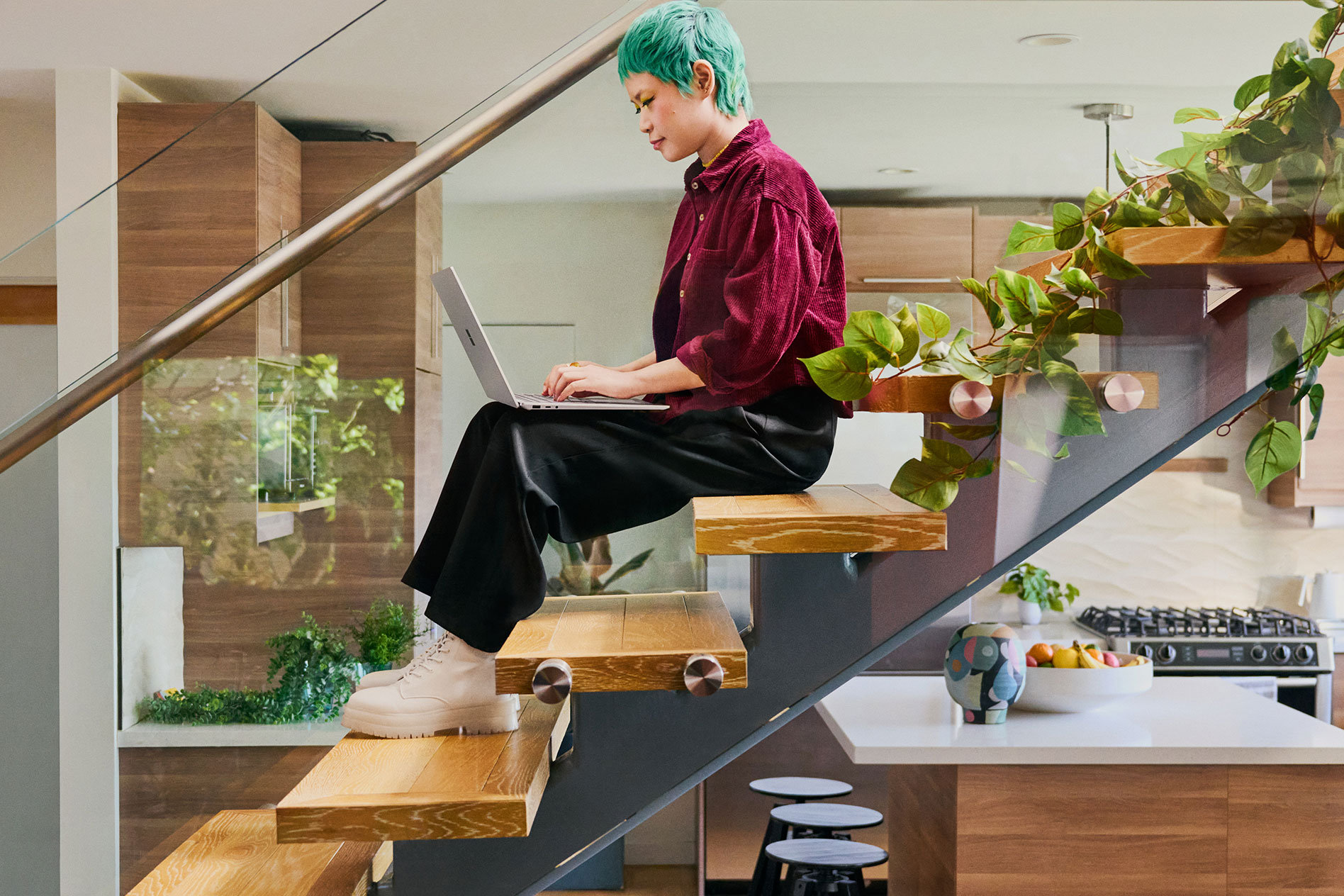 Uma pessoa jovem sentada numa escadaria numa cozinha moderna a escrever no seu Surface Laptop 5.