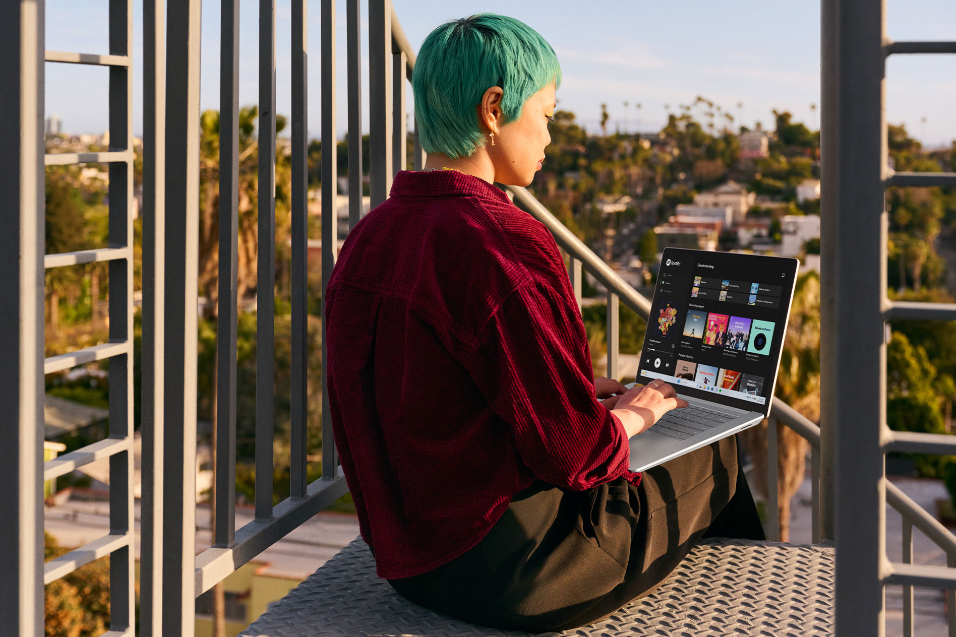 A young person sits in the sunshine on an outdoor metal staircase while typing on their Surface Laptop 5.
