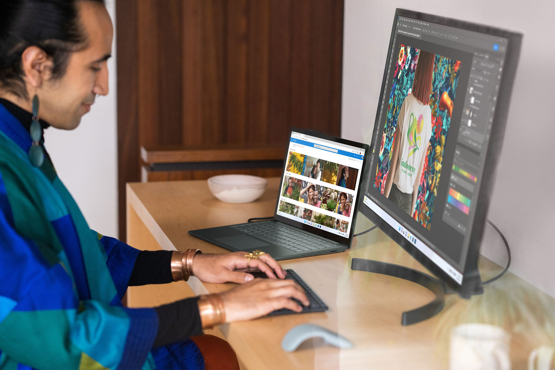 A person sits at a wooden desk typing on an external keyboard with both Surface Laptop 5 and an external monitor showing photos on screen.