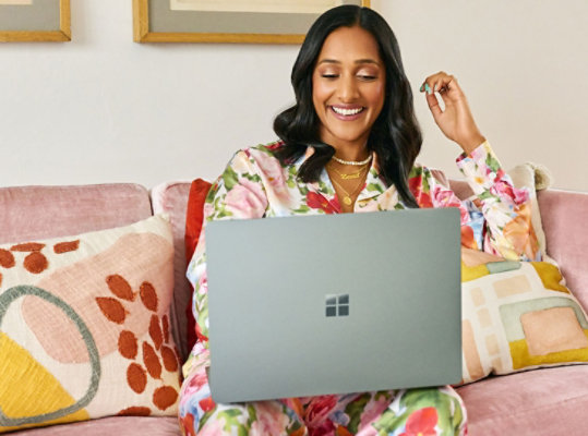 A women wearing colorful clothing sits on a couch with a Surface Laptop 5 on her lap.
