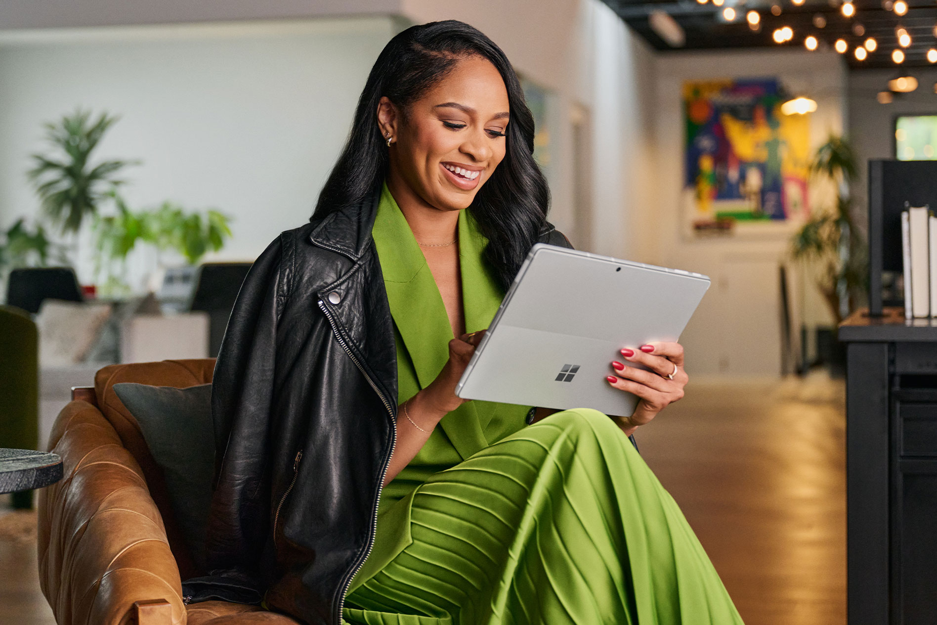A woman wearing a long green dress and black leather jacket sits on a chair interacting with her Surface Pro 9 in tablet mode.