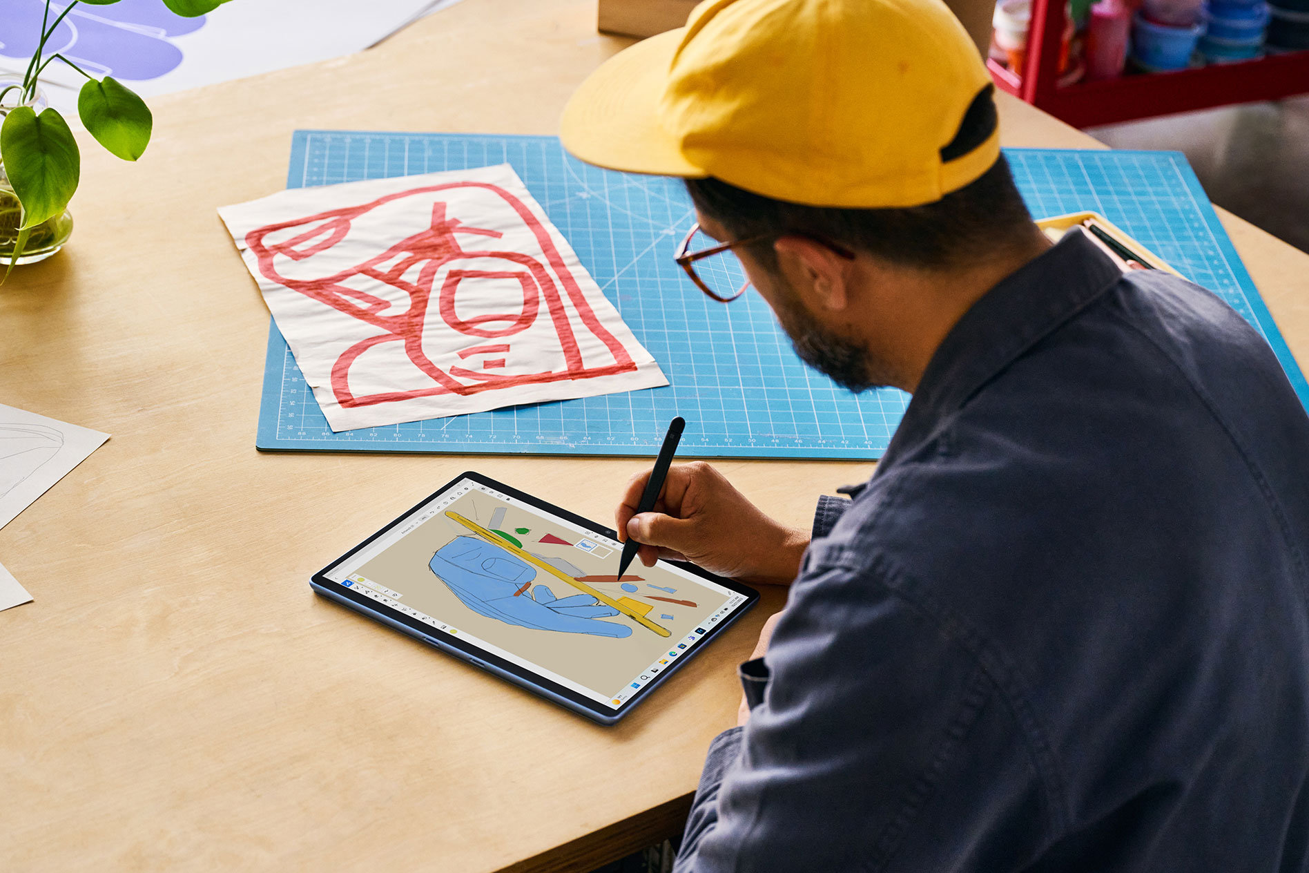 A man wearing a baseball cap sits by a wooden table and draws with a Surface Slim Pen 2 on his Surface Pro 9 in tablet mode.