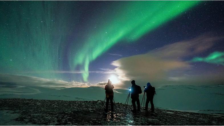 Tre persone con fotocamere su treppiedi che riprendono aurore boreali.