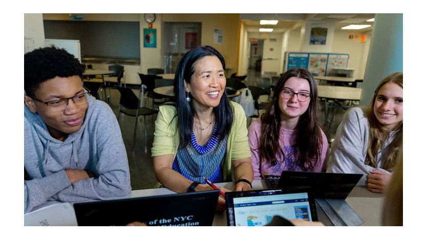 A group of students and a teacher sit at a table with computers.