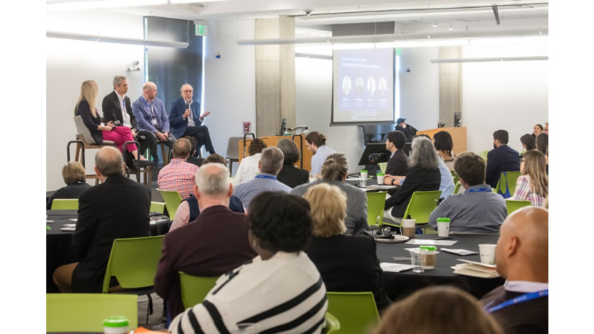 Sign on an easel reading “Washington Local Journalism Summit” in the foreground, with attendees mingling in a bright indoor conference space behind it.