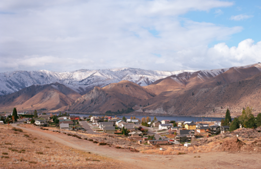 View of a small community next to the Columbia river. The surrounding land is arid; mountains in the background are snow-capped.