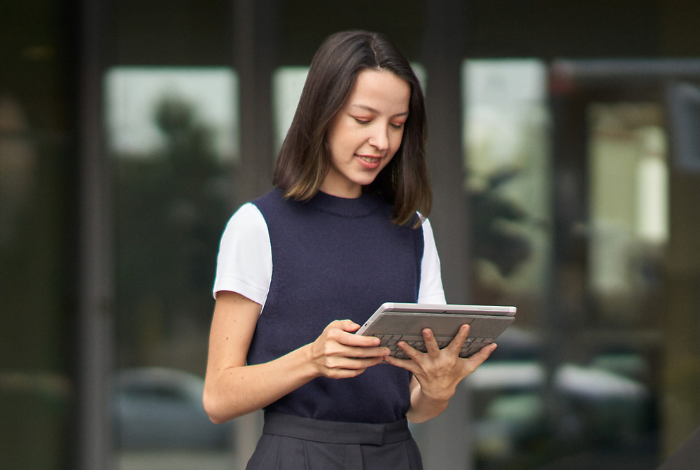 Una mujer está al aire libre, sosteniendo y mirando un dispositivo de tableta. Lleva una camiseta blanca bajo una parte superior sin cubierta. 