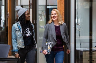 Două persoane mergând pe un hol, stând de vorbă.