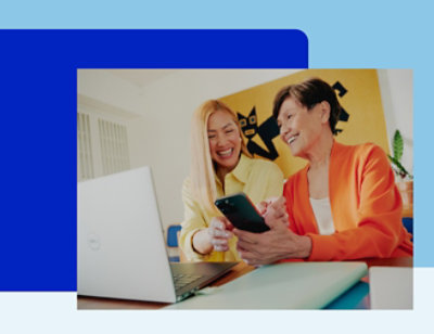 Two women smiling while looking in the phone while a laptop is open on the table