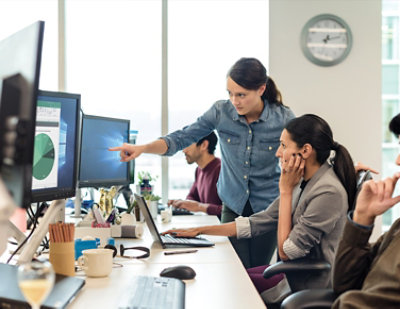 Two women and two men working at desks in an office, with one woman pointing at a computer screen displaying a chart.