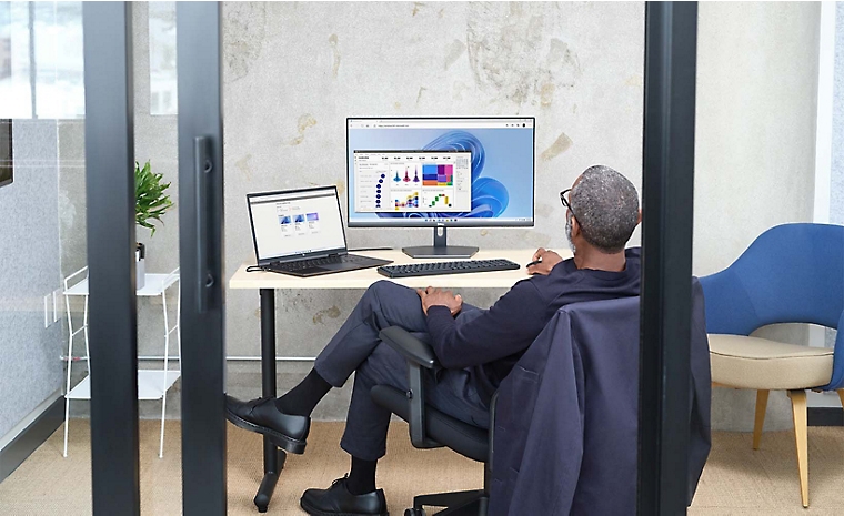 Asmuo, sėdintis biure stikliniais langais, naudoja nešiojamąjį kompiuterį, prijungtą prie stalinio monitoriaus
