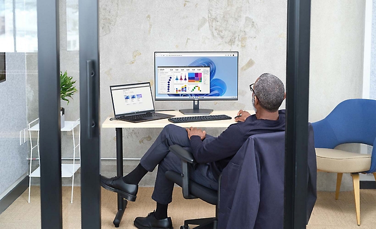 A person sitting in a glass office using a laptop connected to a desktop monitor