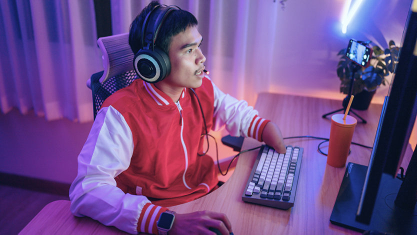 A gamer wearing headphones plays at a keyboard in a purple-lit streaming setup.