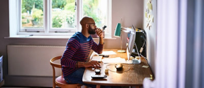 Osoba siedzi przy drewnianym biurku i pracuje na komputerze w pokoju z dużymi oknami.