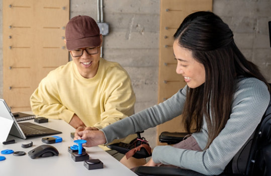A woman who uses a wheelchair testing a selection of Microsoft Adaptive Accessories on Surface Pro with a colleague.