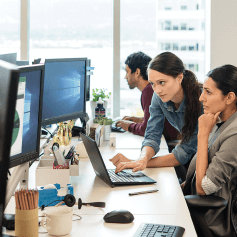 Two people working together at a desk. One is standing and the other is sitting in a desk chair.