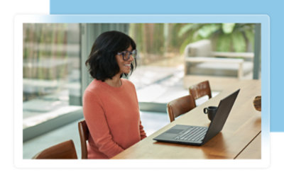 Una donna con un maglione arancione seduta a un tavolo di legno, mentre guarda un laptop in una stanza luminosa.