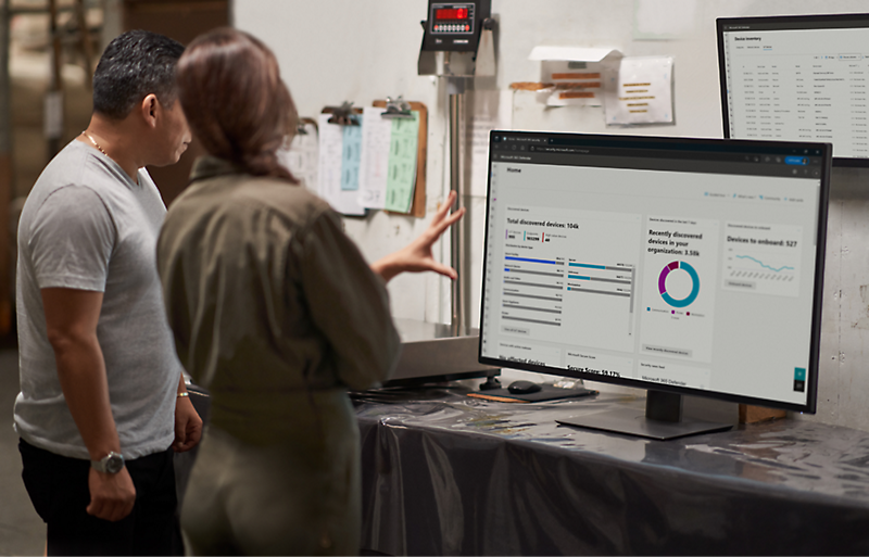 Two individuals stand next to a monitor displaying charts and graphs in a workspace. One person points to the screen