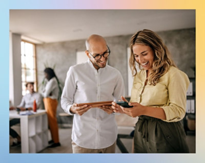 Two individuals standing and interacting with a digital tablet in an office environment with colleagues in the background.