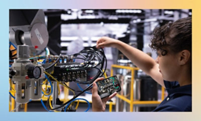 Eine Person schließt in einer industriellen Umgebung Kabel an eine Maschine an und hält dabei ein Bauteil in einer Hand.