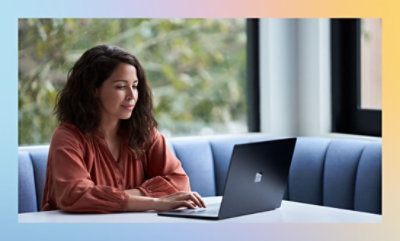 Person in a rust-colored shirt working on a laptop at a table with a blue couch and window in the background.