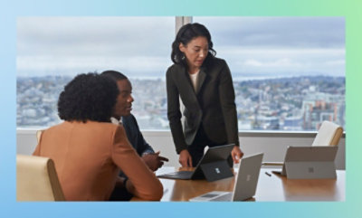 会議室に 3 人の人物がいて、テーブルの上にノート PC が置かれています。1 人が立ち上がってスクリーンを指しています