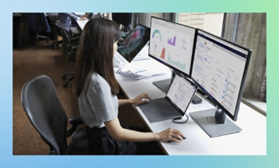Woman working at a desk with multiple computer monitors displaying graphs and data.