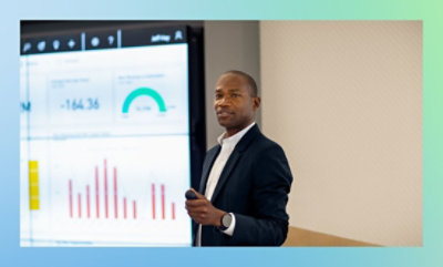 スーツを着た人が、チャートとゲージが表示された大きな画面にデータを提示しています。