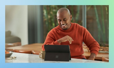 Person in a red sweater sitting at a table with a tablet and stylus, in a room with greenery in the background.